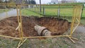 A trench for replacing sewer pipes and other utilities has been dug in a lawn with fallen fall leaves and a dirt path and fenced o