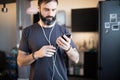 Trendy bearded guy wearing casual grey t-shirt listening music in earphones, checking social networks on smartphone Royalty Free Stock Photo