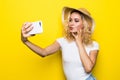 Trendy beautiful cool blond girl wearing a straw hat taking selfie with mobile phone against a yellow wall. Royalty Free Stock Photo