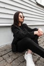 Trendy lovely urban young woman in fashionable sunglasses in stylish youth jacket in shoes enjoy a rest near vintage wooden wall Royalty Free Stock Photo