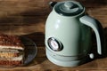 Trendy metal green electric kettle with a thermometer, made in vintage style, stands on a rotating stand next to the cakes. Table