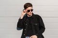 Trendy portrait fashionable young man in stylish denim black jacket in vintage sunglasses with hairstyle on sunny day near white Royalty Free Stock Photo