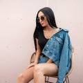 Trendy pretty young hipster woman in stylish sunglasses in a fashionable denim jacket is sitting on a vintage straw chair Royalty Free Stock Photo