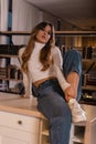 Trendy pretty young woman with red hair in a stylish T-shirt in fashionable blue jeans sitting on a table indoors. Attractive girl