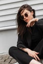 Trendy sexy urban young woman in fashionable round sunglasses in stylish black youth jacket enjoy a rest near vintage wooden wall Royalty Free Stock Photo