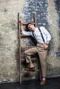Trendy stylish young model man in white shirt and beige pants posing and lean on old wooden ladder. Royalty Free Stock Photo