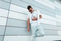 Trendy young hipster man in a fashionable white t-shirt in dark sunglasses in ripped jeans posing near a modern building on the Royalty Free Stock Photo