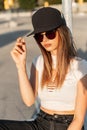 Trendy young hipster woman in fashion sunglasses in stylish youth clothes straightens american black cap on sunny day in parking Royalty Free Stock Photo