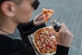 Trendy young man hipster in dark sunglasses with a slice of tasty hot pizza in the street. Handsome guy is sitting in the city Royalty Free Stock Photo