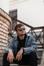 Trendy young man hipster in stylish denim blue clothes trendy in a black sunglasses sits on a vintage staircase Royalty Free Stock Photo