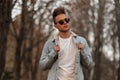 Trendy young man straightens a stylish denim jacket. Handsome hipster guy in black sunglasses with fashionable hairstyle posing in Royalty Free Stock Photo