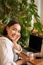 Trendy young woman sitting in cafe and smiling at camera, drinking coffee and using laptop, working remotely, studying Royalty Free Stock Photo