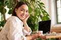 Trendy young woman sitting in cafe and smiling at camera, drinking coffee and using laptop, working remotely, studying Royalty Free Stock Photo
