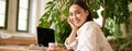 Trendy young woman sitting in cafe and smiling at camera, drinking coffee and using laptop, working remotely, studying Royalty Free Stock Photo