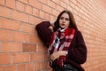 Trendy young woman in a warm knitted sweater with a stylish scarf in leather pants is resting near a vintage brick wall in the Royalty Free Stock Photo