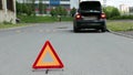 Triangle warning sign on road foreground and broken car with blinker lights on road wayside