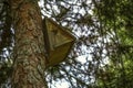 A triangular birdhouse with a round entrance and a peg for resting birds on a coniferous tree.