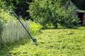 a trimer lawn mower stands by a fence on a mowed lawn near a house in a rural area.