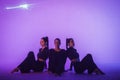 Trio of young acrobats posing sitting sitting on floor in studio with purple light. Gymnast girls in black costumes