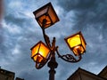 triple lantern 3 head with transparent glass panels and electric bulb, iron street lighting glowing in evening against Royalty Free Stock Photo