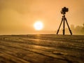 Tripod photo camera on wooden bridge films bright sunrise through over lake and trees very thick golden fog