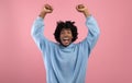 Triumphant black teen guy lifting arms above his head, shouting in excitement on pink studio background