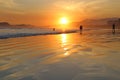 Sea with small waves, sand on the beach, mountains in the background, clouds from the sky, the sun sets, reflections in water and