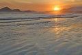 Sea with small waves, sand on the beach, mountains in the background, clouds from the sky, the sun sets, reflections in water and