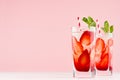 Tropical refreshing fruit drink with slices strawberry, green mint, ice, straw on soft light white wood board, pastel pink.