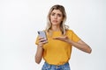Troubled young woman asking help with mobile phone, pointing at smartphone and looking upset, standing over white Royalty Free Stock Photo