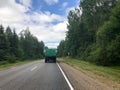 A truck, a tractor with a large green trailer is driving along a forest asphalt road with green trees on the grounds Royalty Free Stock Photo