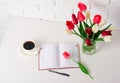 Tulip flowers are in a vase on the table, cup of coffee and diary, white brick wall as background