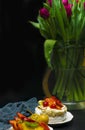 Tulip in vase with cup of coffee and fruit cake on dark background. Coffee time. Flower gift. Pavlova pie