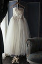 A tulle and embroidered lace wedding dress hanging on a screen with vintage shoes and an embroidered chaise