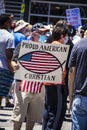 Older man in stars and stripes hat with Covid-mask hold up sign reading Proud American Christian in crowd