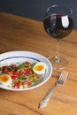 Tuna salad with vegetables, boiled eggs, cherry tomatoes and spices and one glass of red wine over wooden background Royalty Free Stock Photo