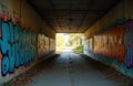Tunnel with concrete walls covered in colorful graffiti art. Paved pathway leads through underpass towards bright, tree-lined Royalty Free Stock Photo