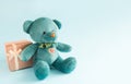 Turquoise soft teddy bear with an embroidered heart holds a gift box and a bow on a blue background. Children`s toy. Love, a gift