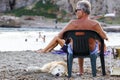 TUSCANY , ITALY 17 JULY 2018 : Mature man resting on a deck chair listening to music petting his dog on the beach