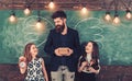 Tutor and pupils smile with school supplies. Happy teacher and schoolgirls at chalkboard. Bearded man and small girls at Royalty Free Stock Photo