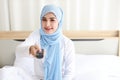 TV and happiness concept. Beautiful asian muslim woman in white sleepwear sitting on bed, holding television remote and watching Royalty Free Stock Photo