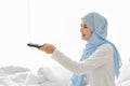 TV and happiness concept. Beautiful asian muslim woman in white sleepwear sitting on bed, holding television remote and watching Royalty Free Stock Photo