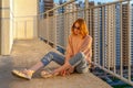 Tween redhead girl in pullover, jeans and sneakers sitting on balcony against high-rise multi-storey residential building at Royalty Free Stock Photo