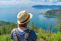 Tween tourist girl in hat and backpack standing on cliff top and admiring beautiful landscape of Baikal lake and Angara river