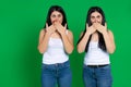 Twin girls in identical clothes covers mouths with both hands being surprised on a green background