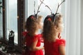 Twin Little Girls Sisters In Red Dresses Staying Near The Window Waiting Santa. Cute Kids With Deer Horns