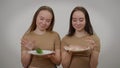 Twin sisters posing with raw meat and fresh organic cucumber at grey background. Portrait of confident meat-eater and