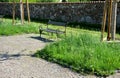 Stone wall in front of it stands a bench of metal twisted pipes and wood on a paved stone path meadow with flowers in a small town