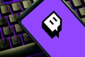 Twitch icon on a cellphone screen on a purple illuminated gaming keyboard. Video game streaming site violet logo