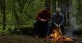 Two adult tourists men are warming near campfire in forest, chatting sitting on log Royalty Free Stock Photo